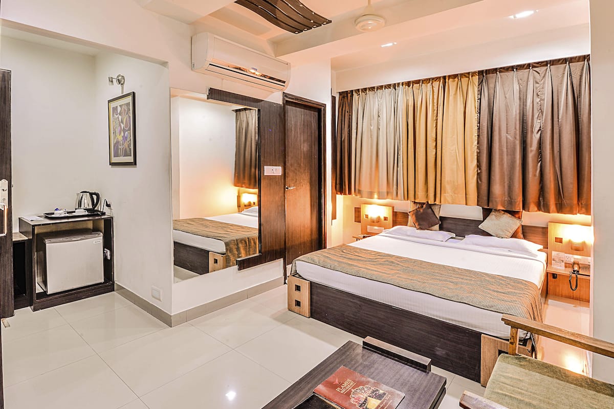 A hotel room with a bed, bedside table, lamp, and a small desk with a refrigerator.