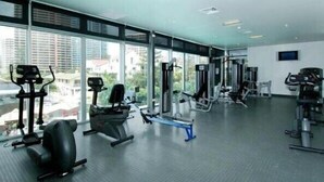 A gym with various exercise equipment, large windows, and a view of buildings outside. - Private Apartments at Q1 (Surfers Paradise)
