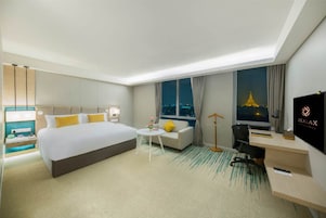 Habitación de hotel con una cama grande, un escritorio, un sofá, una mesita con flores, un televisor y vistas a la ciudad de noche.