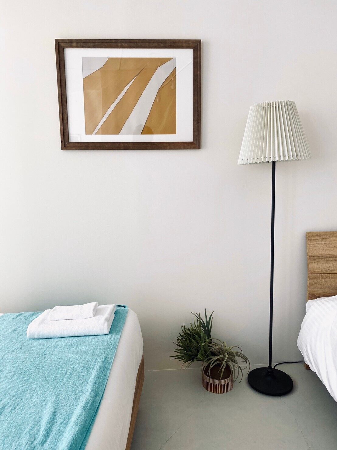 Una cama bien hecha con una manta azul, un cuadro abstracto enmarcado en la pared, una lámpara de pie alta y una pequeña planta en maceta sobre el suelo.