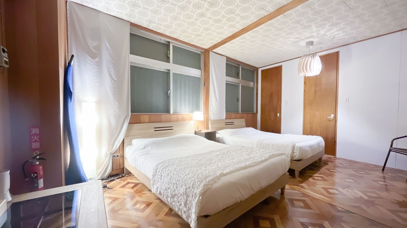Habitació d'hotel amb dos llits, un sòl de fusta, una finestra gran amb cortines, un extintor i una porta.