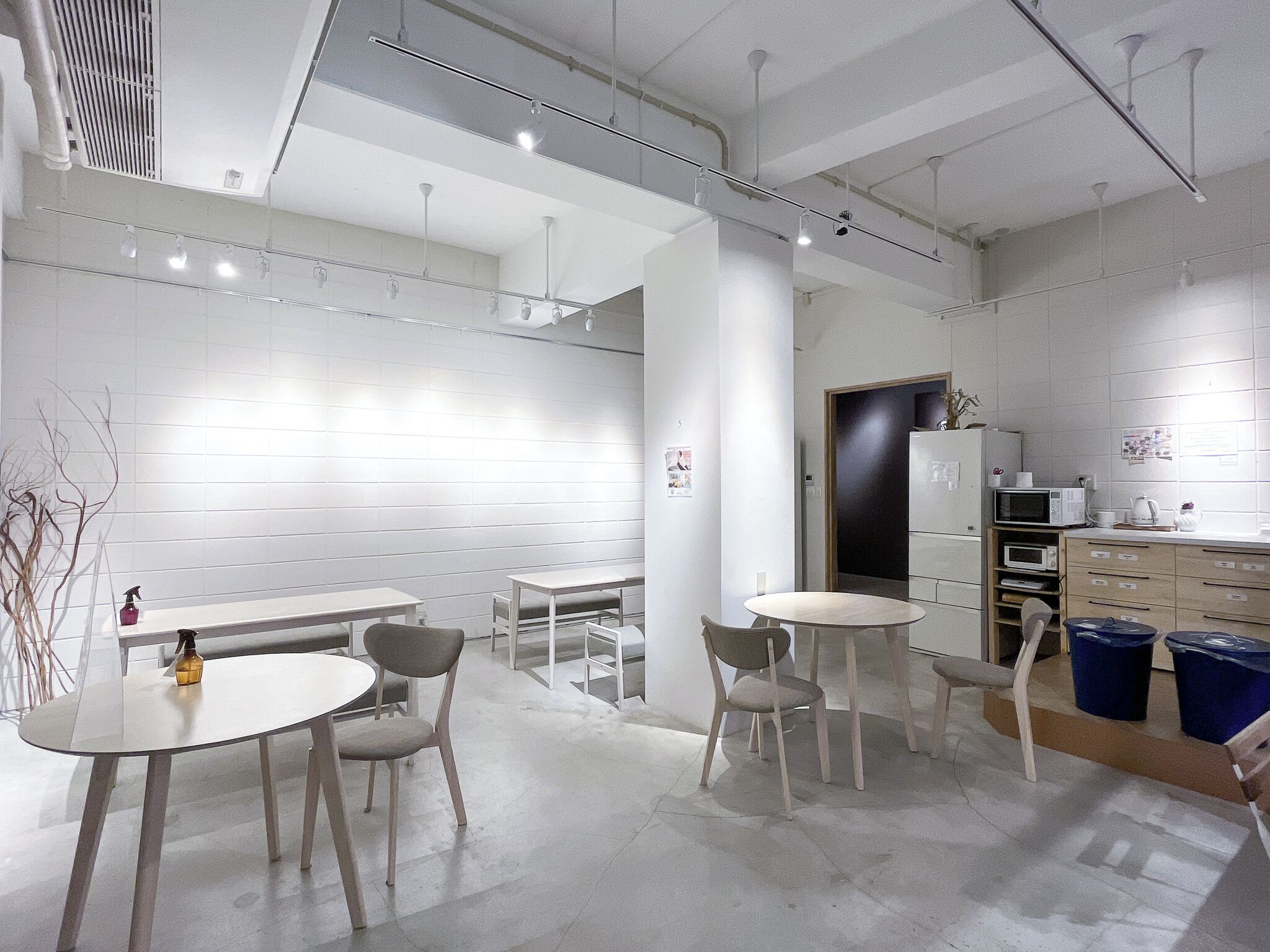 A modern, well-lit room with white walls, a large window, and a kitchen area with a microwave and cabinets. There are several tables and chairs arranged for dining or working.