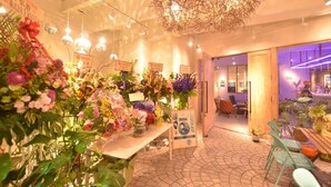 A well-lit indoor space with a variety of potted plants and flowers arranged on tables, a wooden bench, and a seating area with chairs. - Lestel Naha - Hostel (Naha)