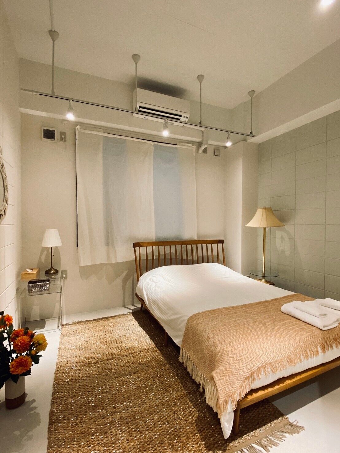 Une chambre moderne avec un lit en bois, une table de chevet avec une lampe, une petite table de chevet avec une boîte de mouchoirs, un vase décoratif avec des fleurs et un tapis à motifs.