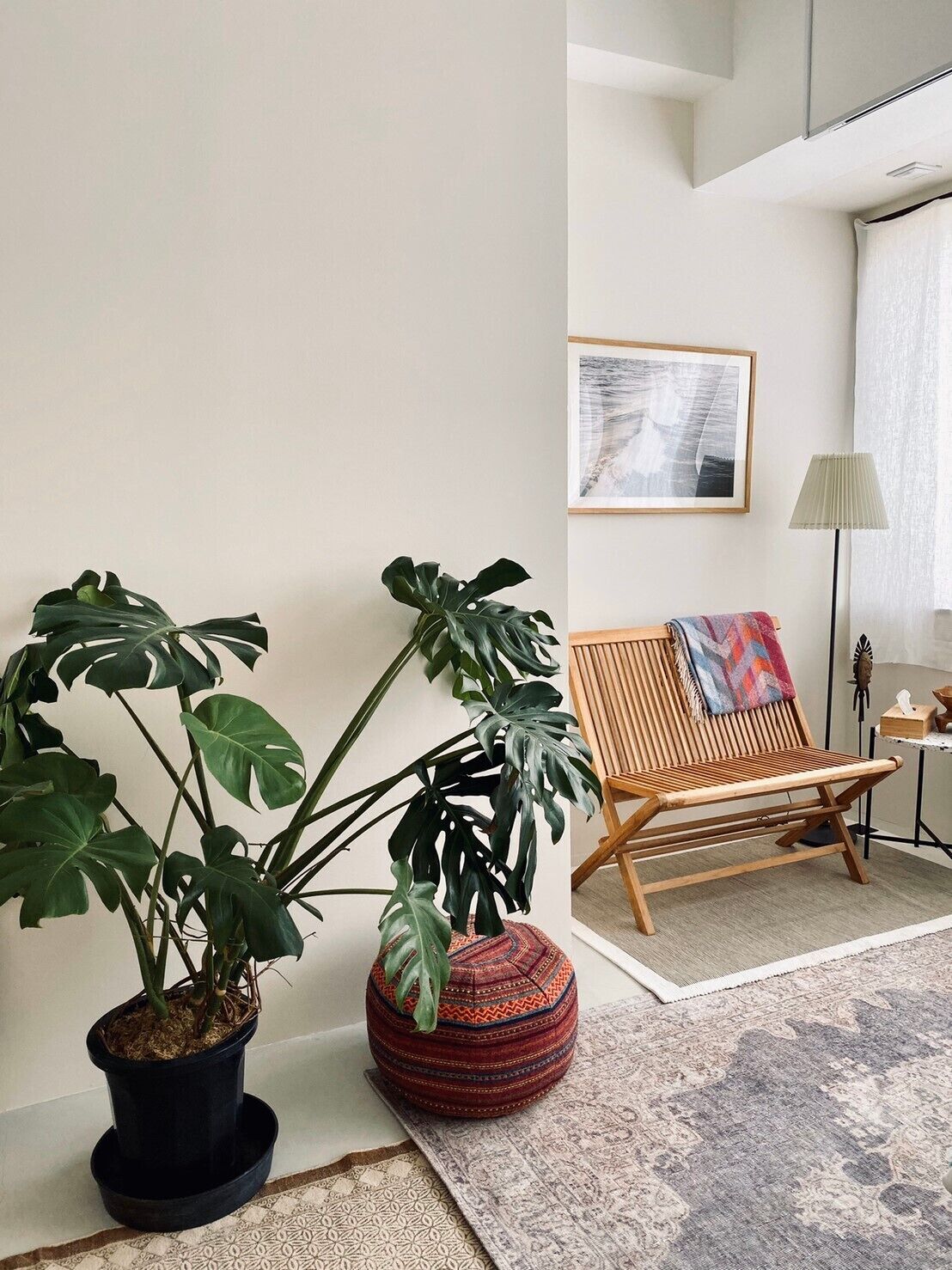 Una habitación con una gran planta de monstera en maceta, un sillón de madera con una manta de colores y un cuadro abstracto enmarcado en la pared.