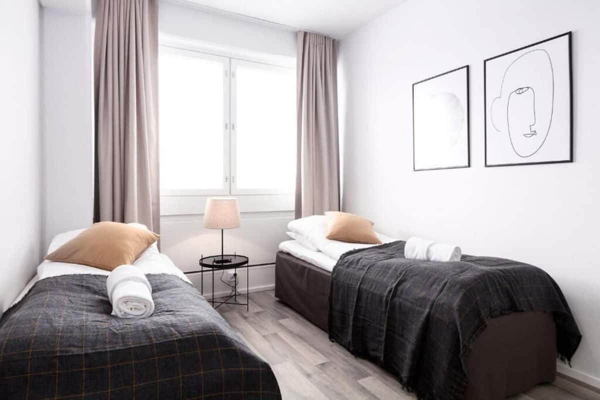 A hotel room with two beds, a window with curtains, a bedside table with a lamp, and two framed pictures on the wall.
