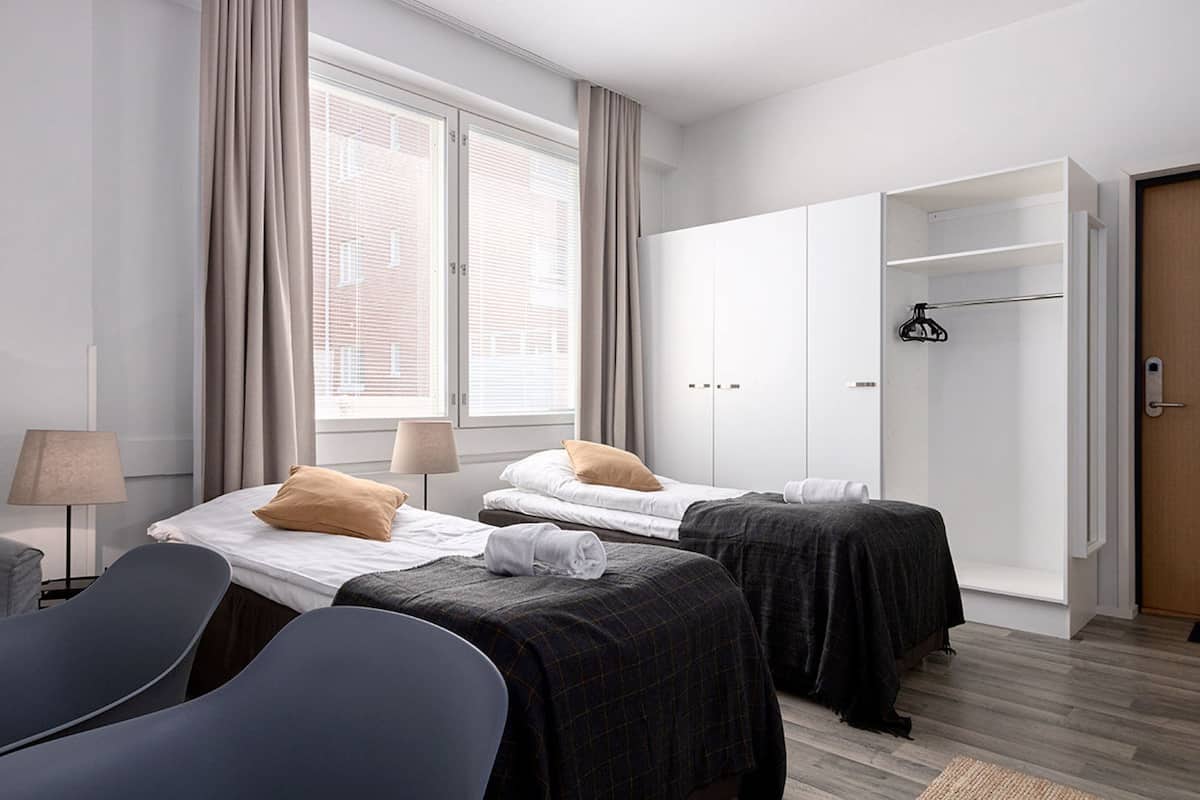A hotel room with two beds, a wardrobe, a chair, and a window with curtains.