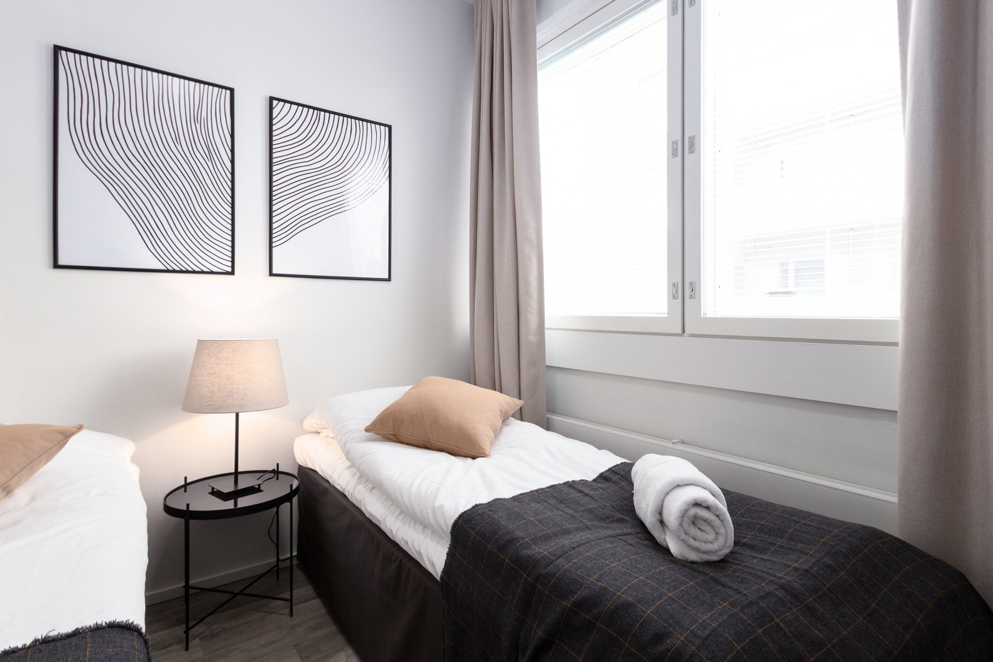Hotellihuone, jossa on kaksi sänkyä, yöpöytä lampun kanssa ja kaksi abstraktia taideteosta seinällä.