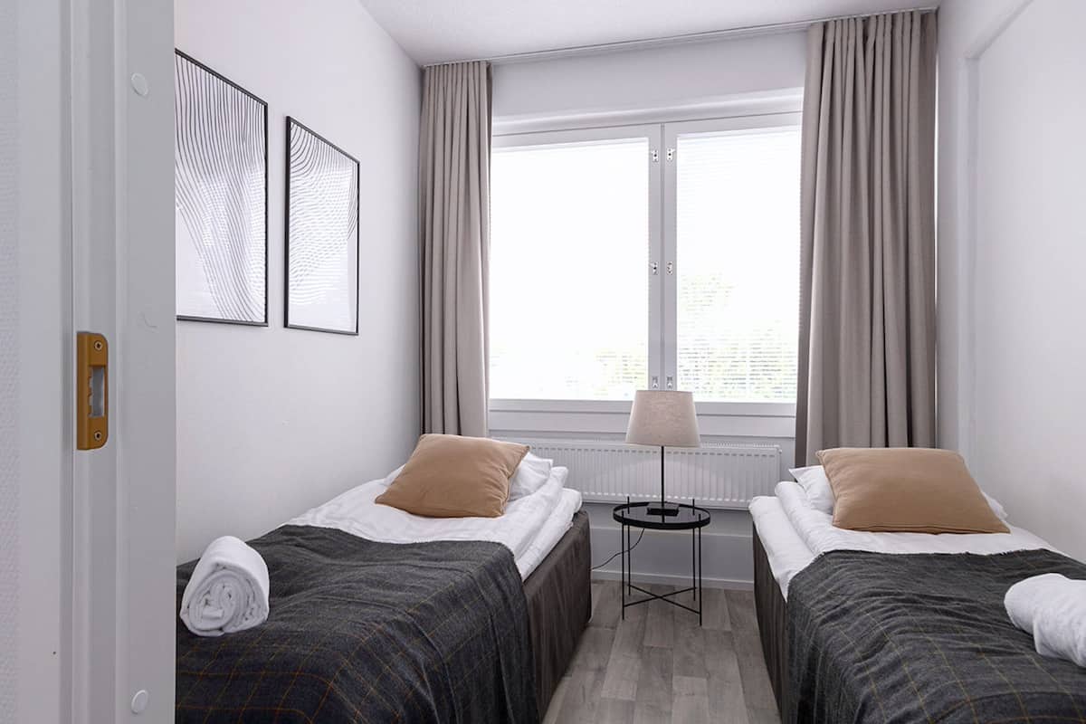 A hotel room with two beds, a small table with a lamp, and a window with curtains.