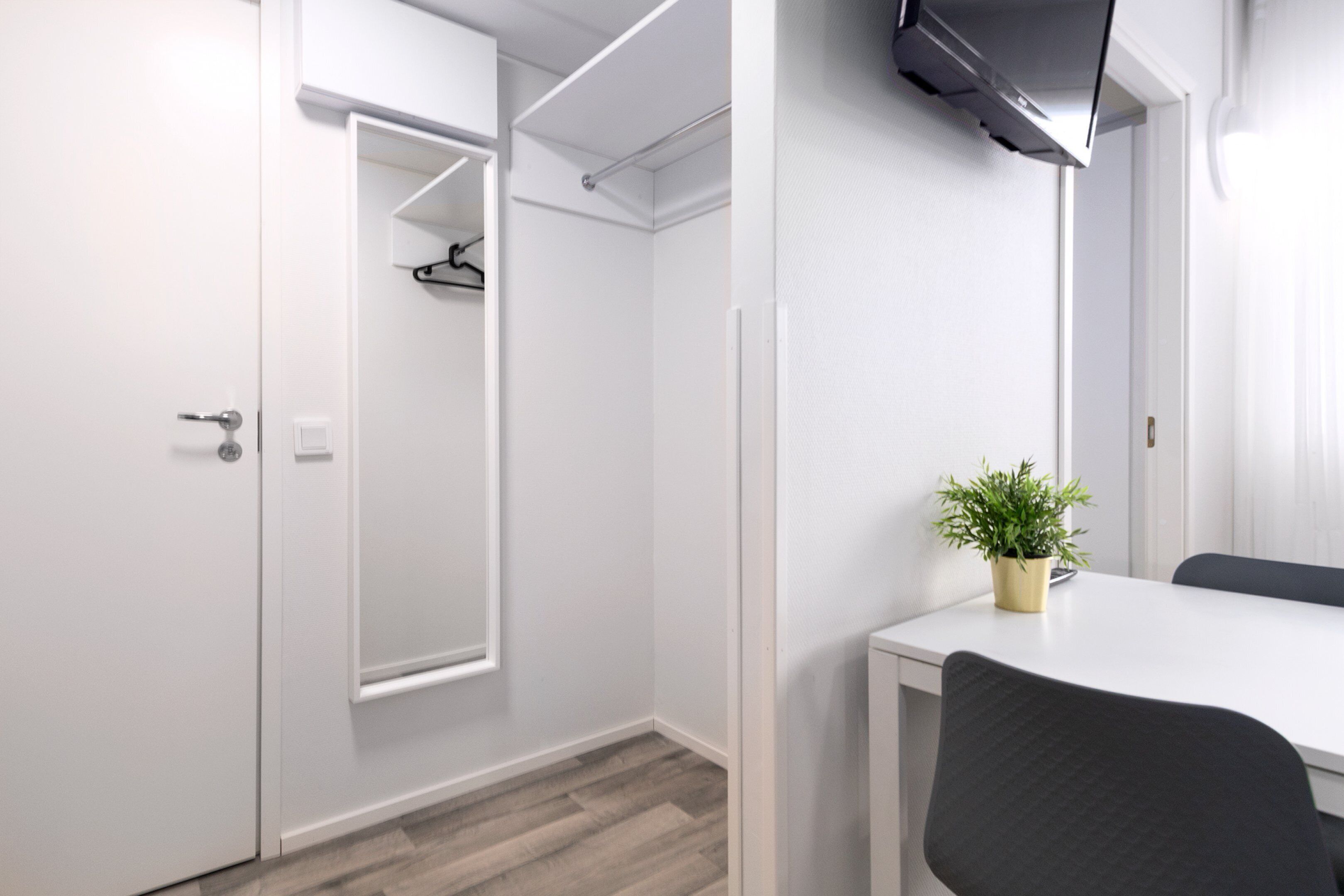 A modern, minimalist room with a white wardrobe, a mirror, a small table with a potted plant, and a black chair.