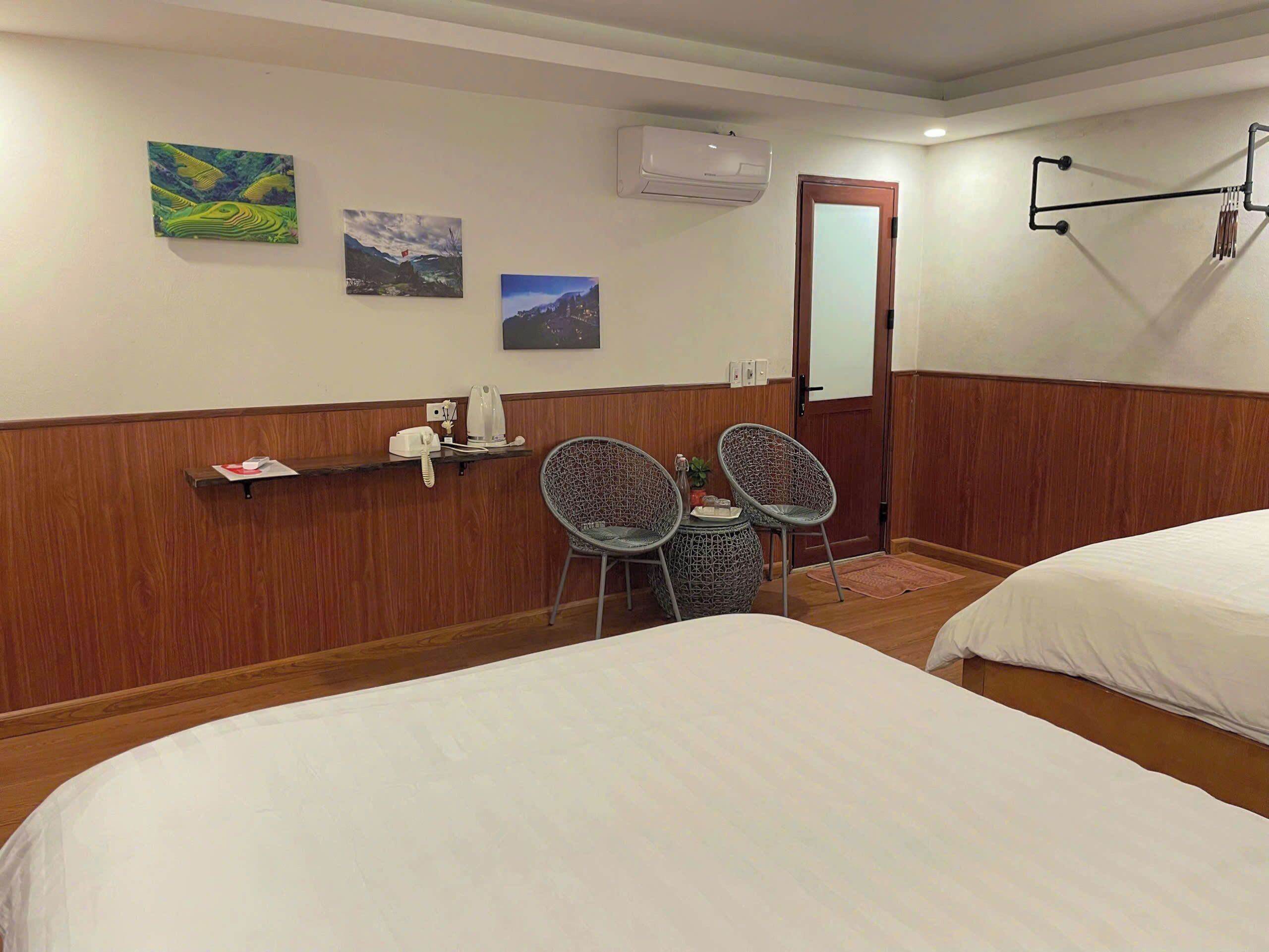 Une chambre d’hôtel avec deux lits, une petite table avec des chaises, une étagère avec un téléphone et une bouilloire, et trois tableaux accrochés au mur.