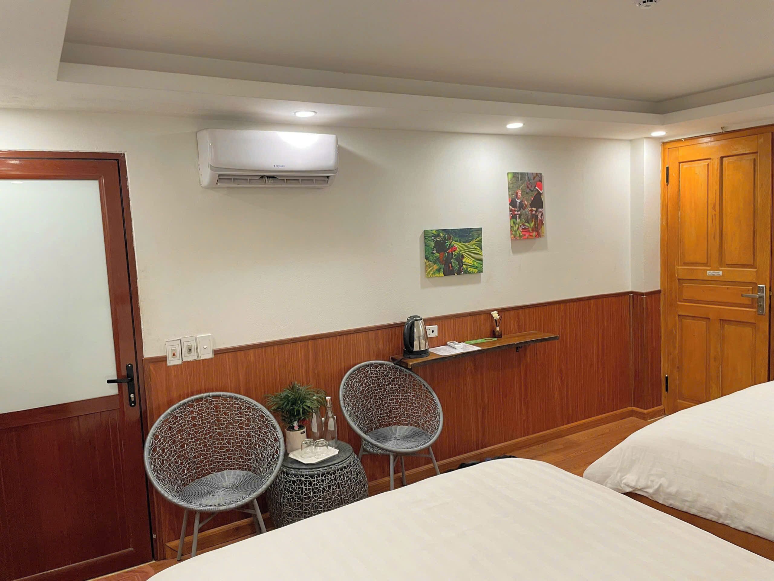 Una habitación de hotel con dos camas, una mesita con hervidor, dos sillas de mimbre, un aire acondicionado y dos cuadros en la pared.