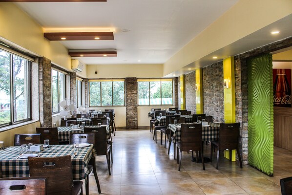A spacious restaurant with multiple tables and chairs, large windows, and a stone wall with a Coca-Cola sign. - La Fortuna Resort and Spa (Mahabaleshwar)