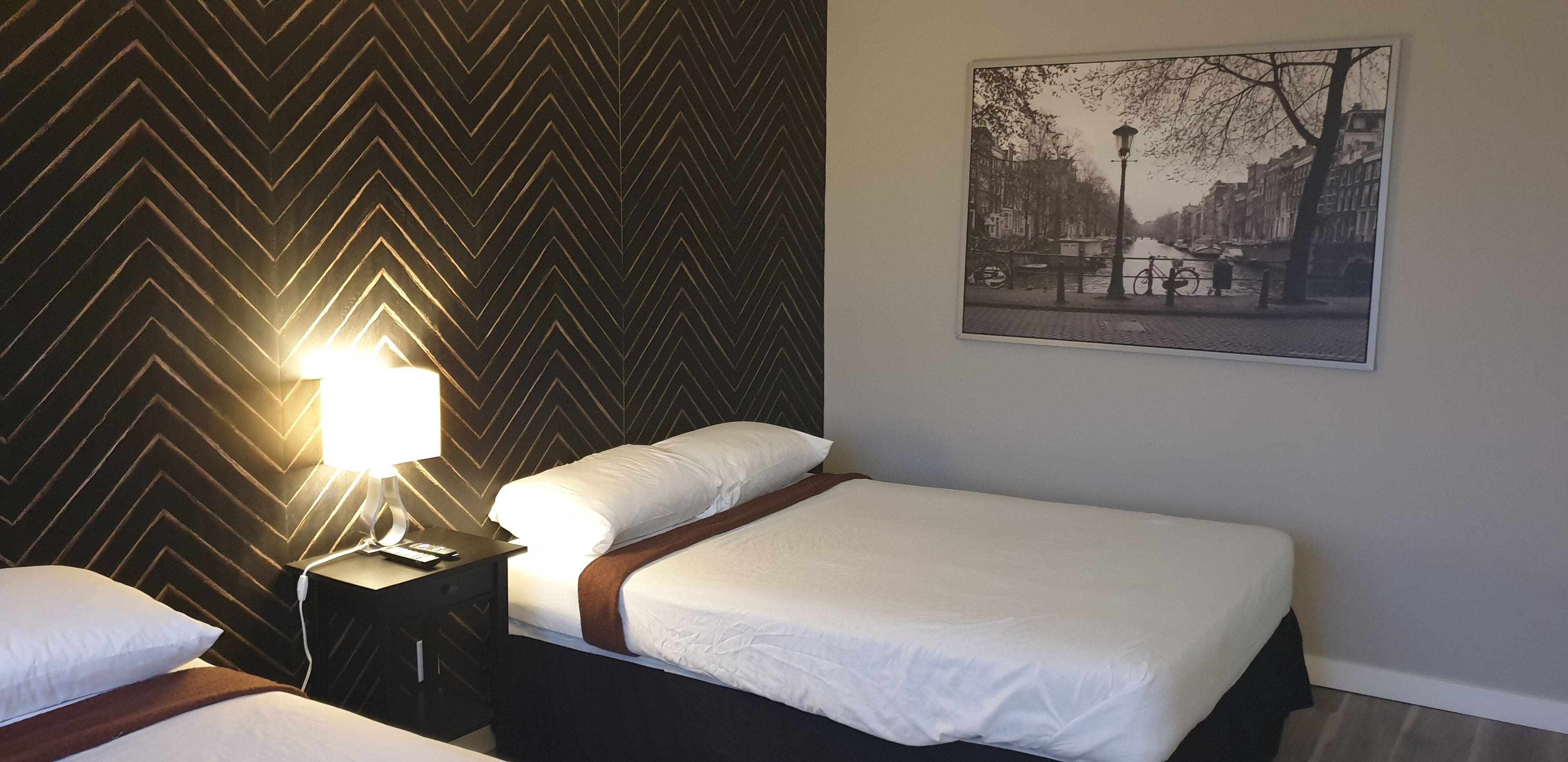 A hotel room with a bed, a nightstand, a lamp, and a cityscape painting on the wall.
