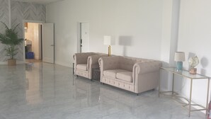 Ein geräumiges, modernes Zimmer mit einem polierten Marmorboden, einer beigefarbenen Sofagarnitur, einem kleinen Tisch mit einer Lampe und einem Blumentopf.