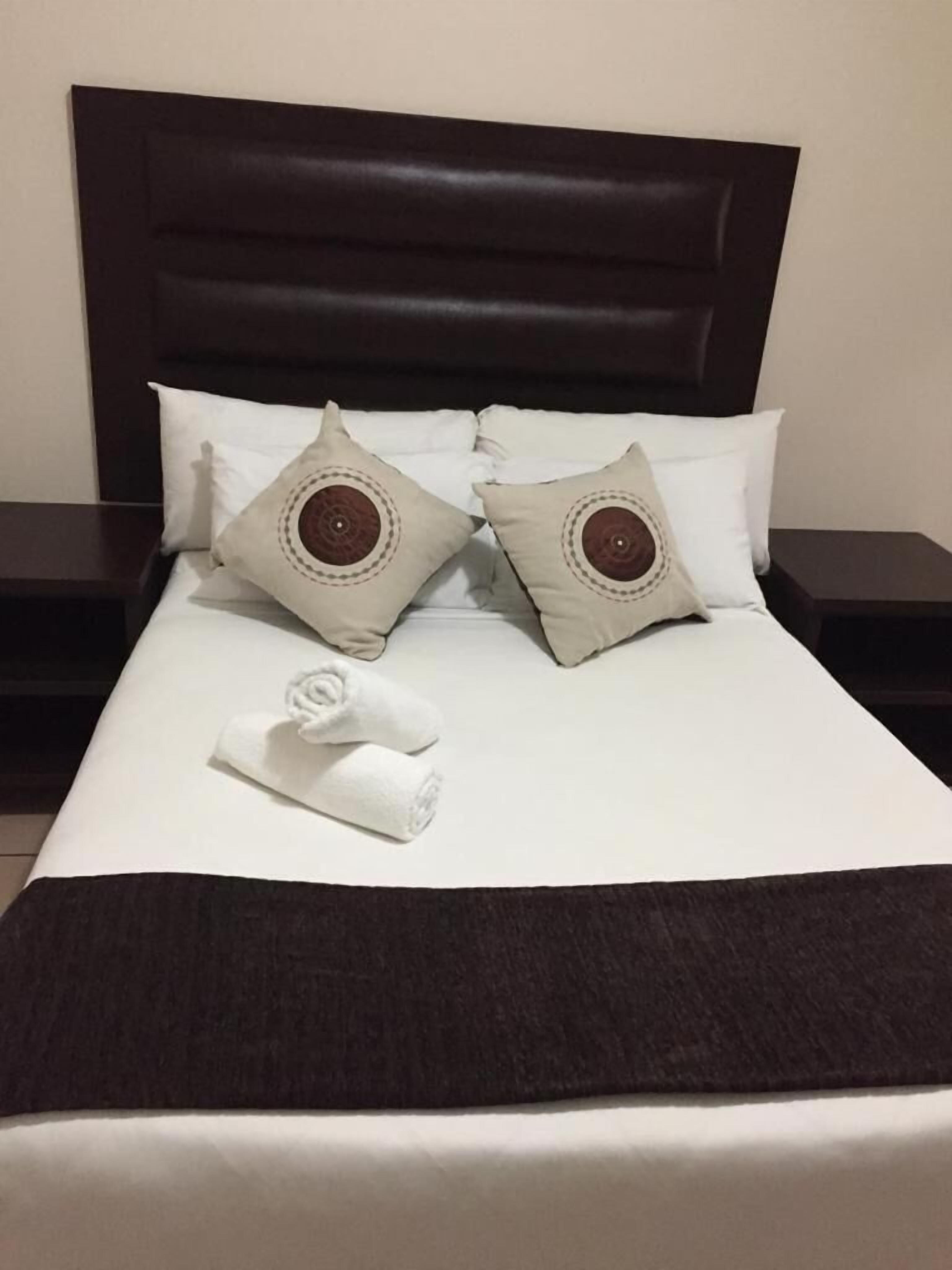 Una cama bien hecha con un cabecero oscuro, dos almohadas decorativas y una toalla doblada.