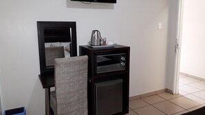 Una habitación pequeña con una cama, un televisor montado en la pared, una kitchenette con cafetera y un basurero.