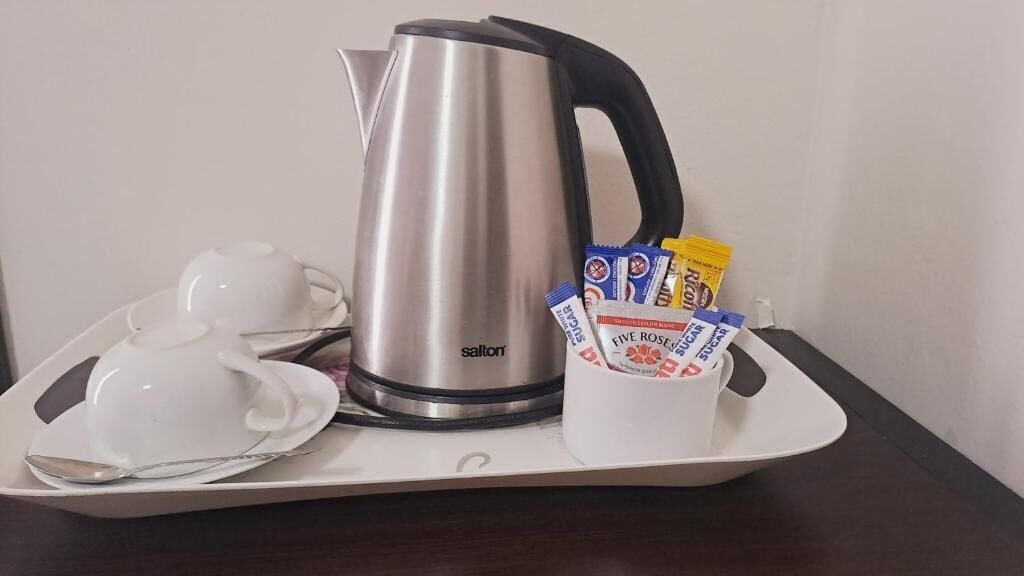 Una tetera de acero inoxidable, una tetera blanca, una taza blanca con bolsitas de té y una bandeja blanca con una cuchara sobre una mesa de madera oscura.