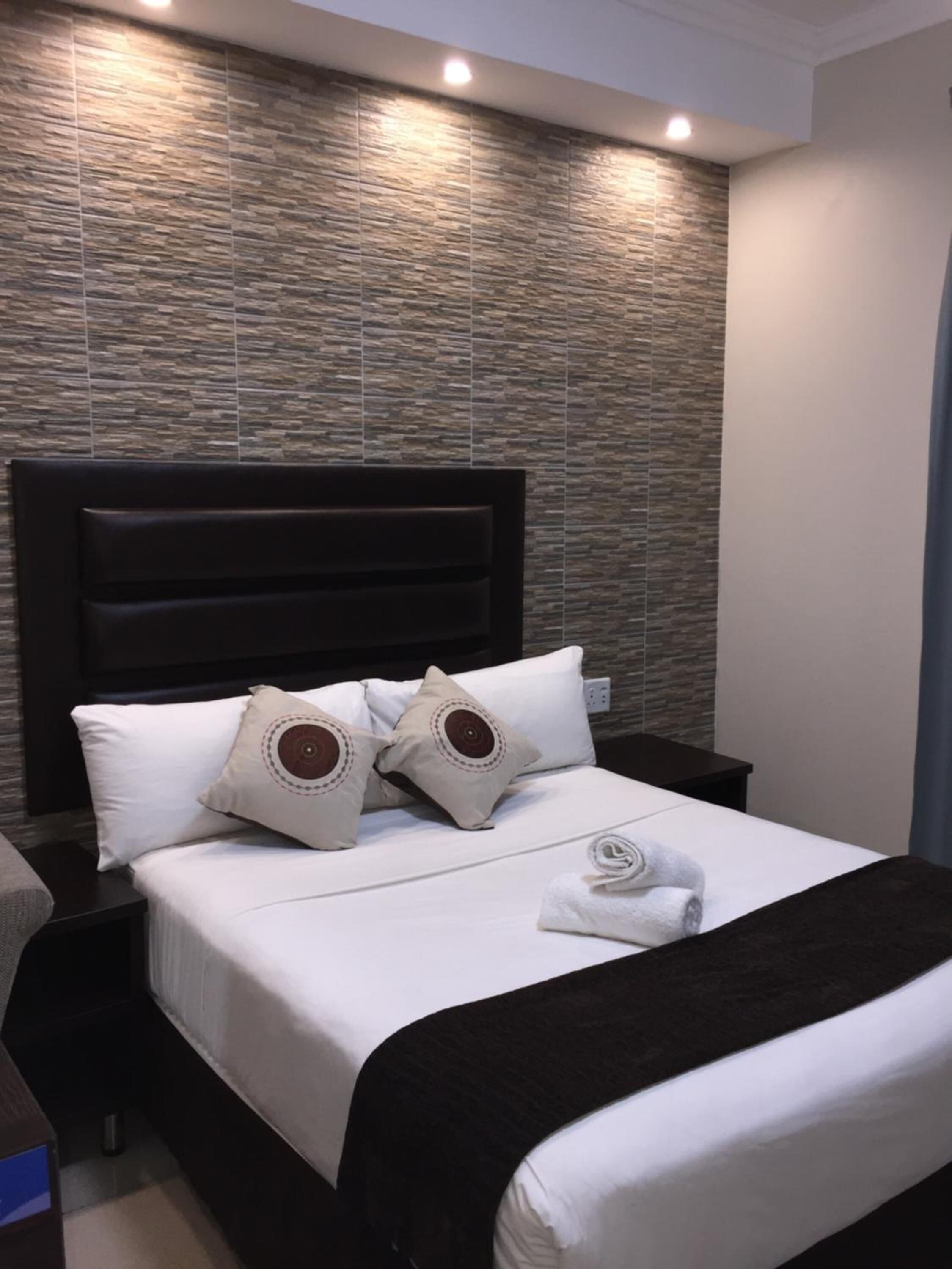 A neatly made bed with white linens and two decorative pillows, a dark headboard, and a wall with a textured pattern.