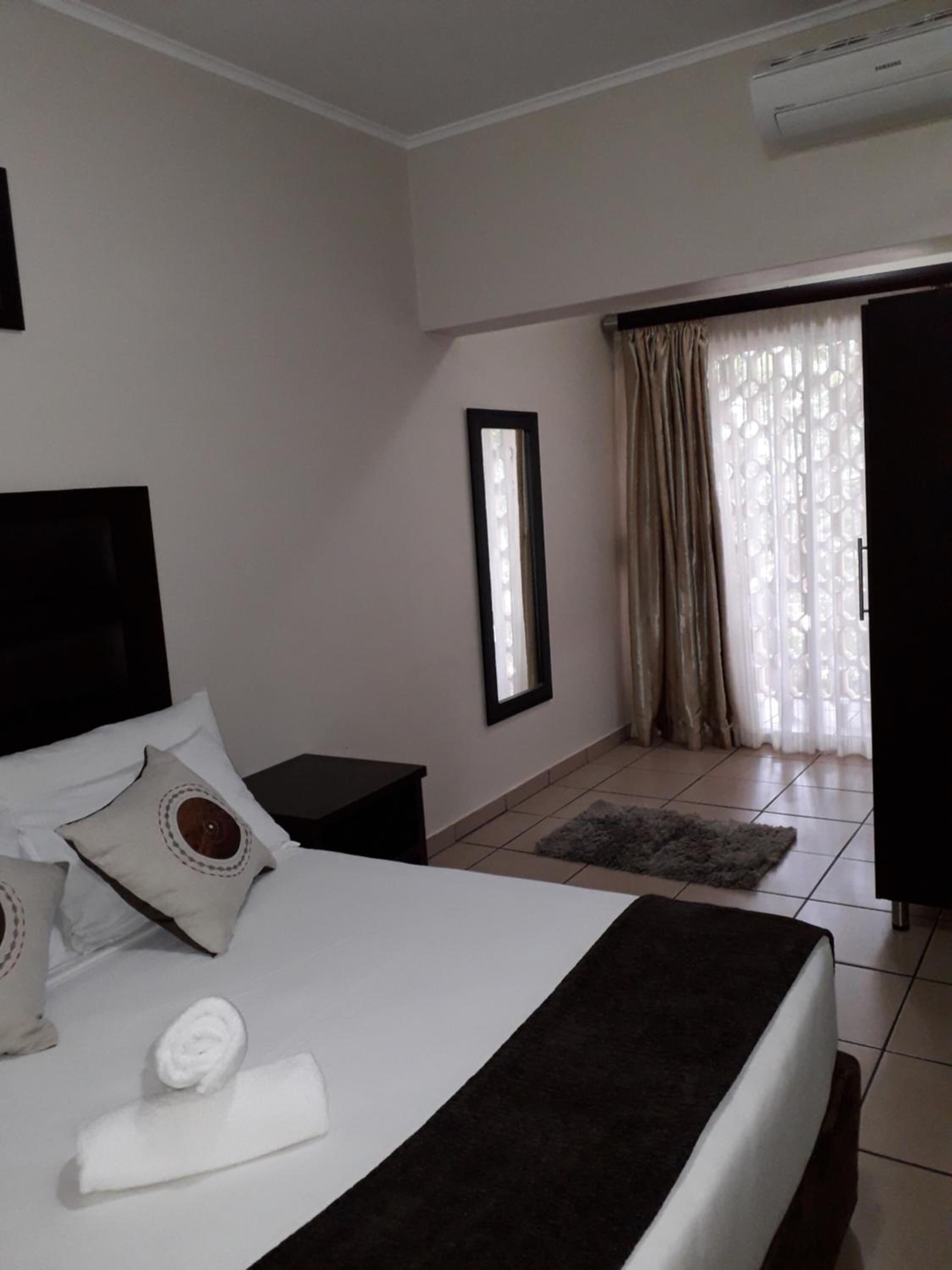 A hotel room with a bed, a bedside table, a nightstand, a window with curtains, and an air conditioning unit.