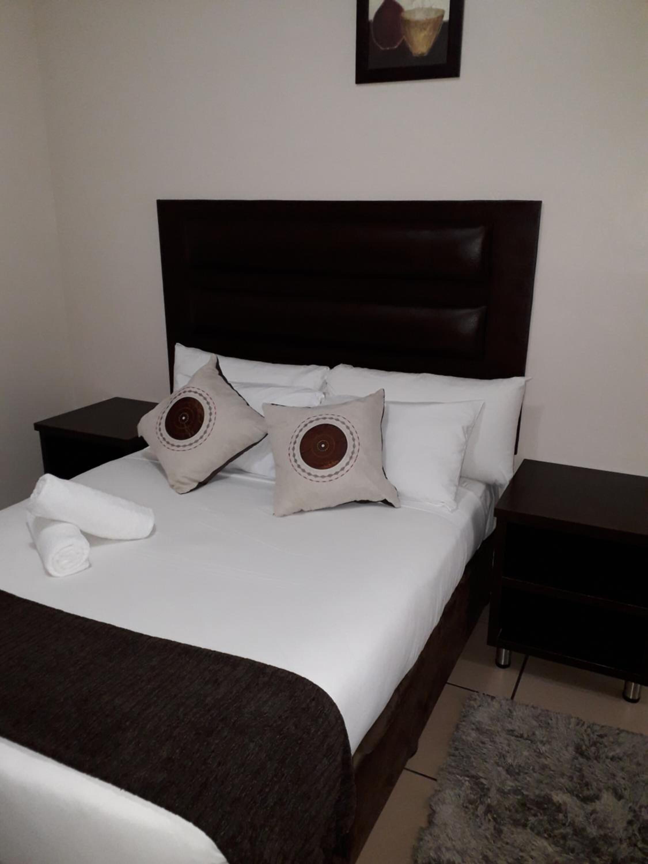 A neatly made bed with white linens, two decorative pillows, a folded towel, and a dark headboard.