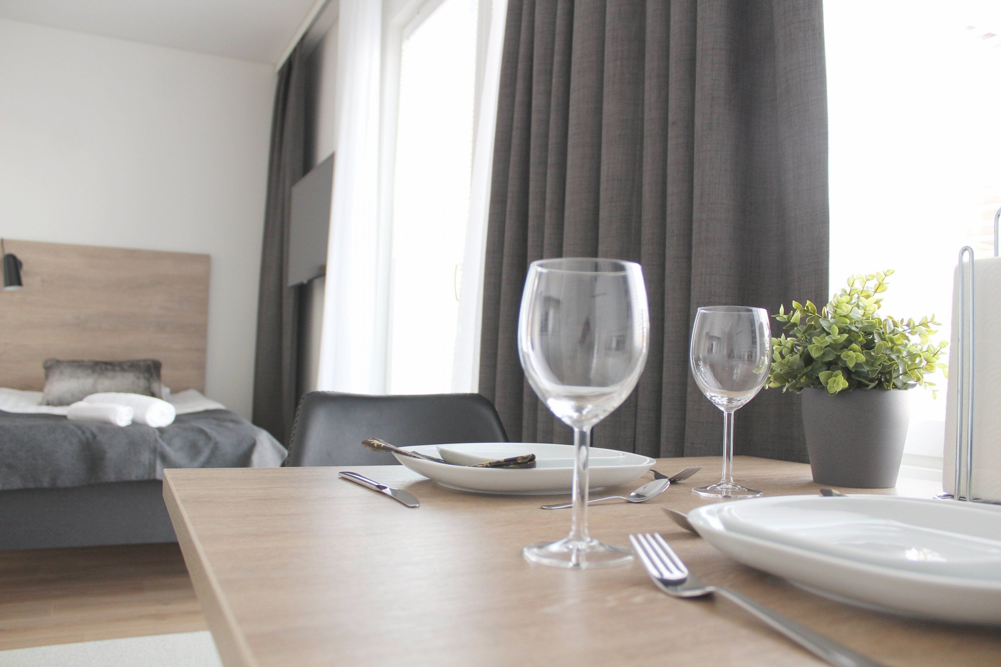 Una mesa de comedor ordenada con un plato, dos copas de vino, cubiertos y una planta en maceta en una habitación de hotel moderna.