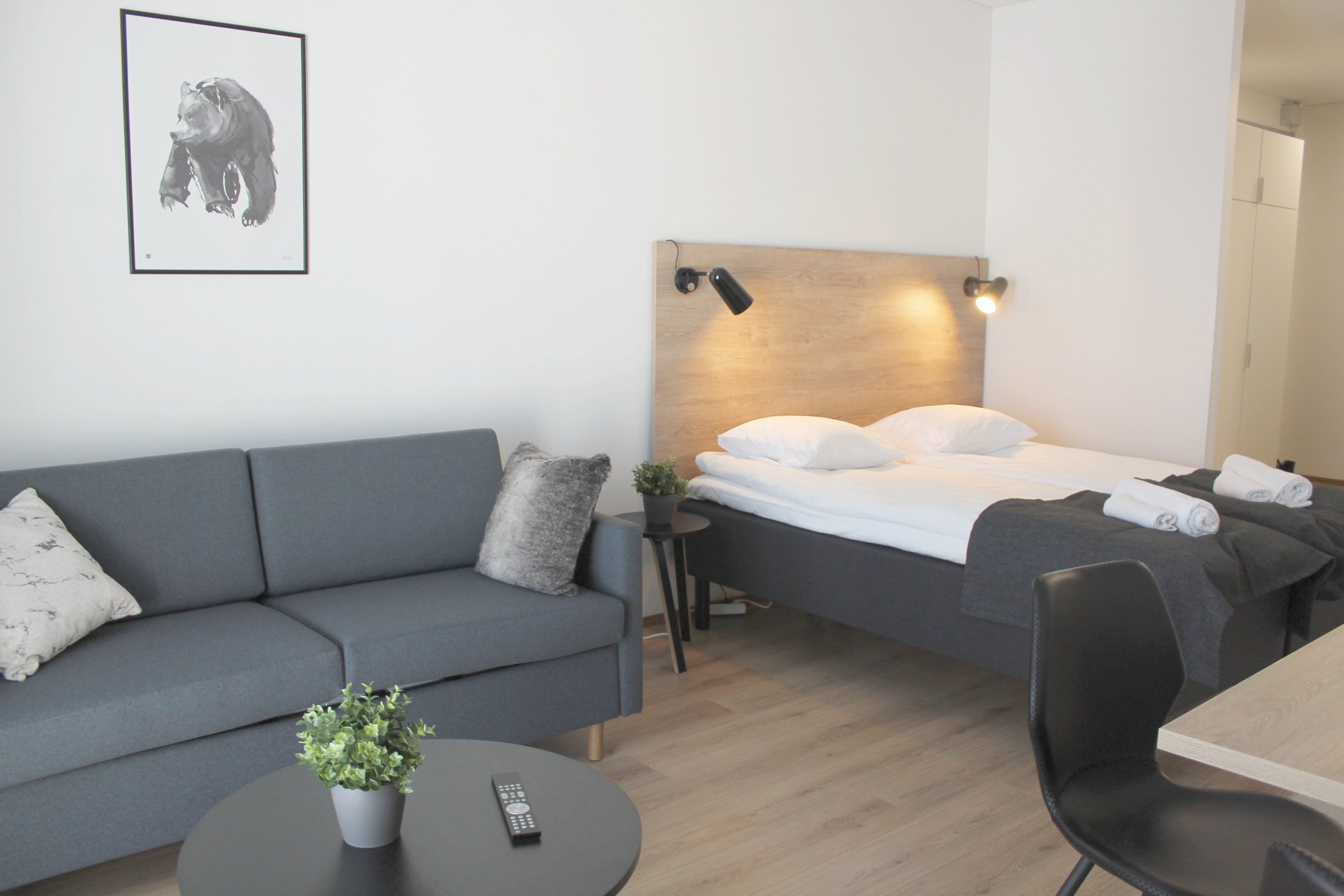 Ein modernes Hotelzimmer mit einer grauen Couch, einem Bett mit weißer Bettwäsche, einem kleinen Tisch mit einer Pflanze und einer Fernbedienung auf dem Tisch.