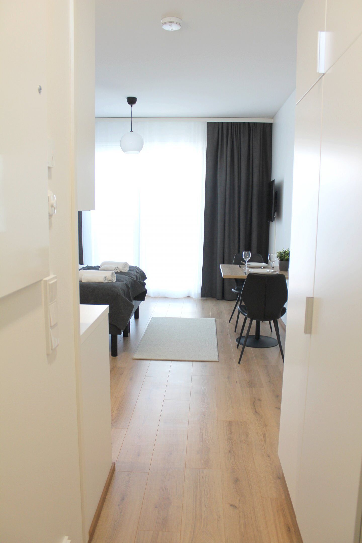 A modern hotel room with a bed, a desk with a chair, a dining table with chairs, a television mounted on the wall, and a large window with curtains.