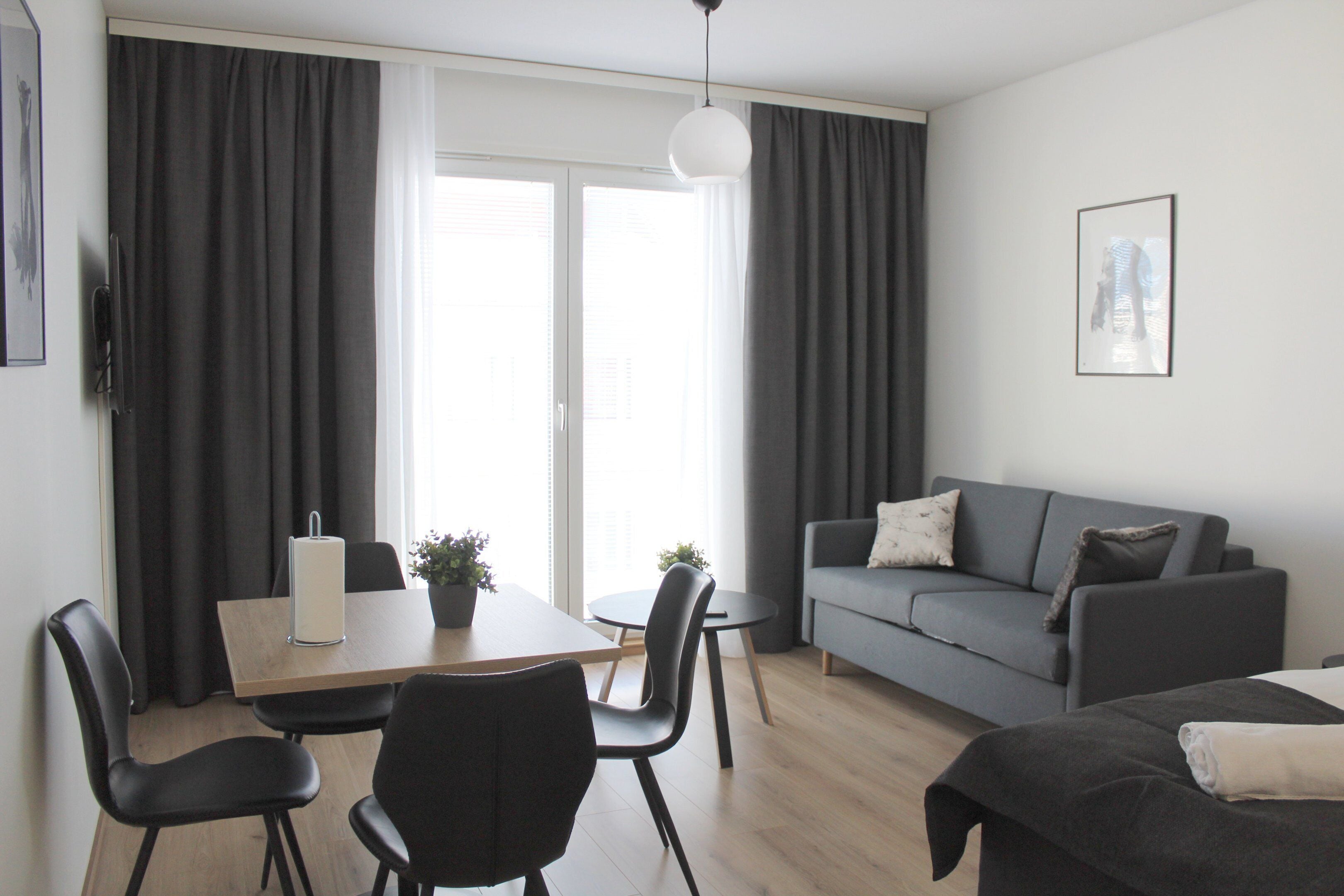 Una sala de estar moderna con un sofá gris, una mesa de comedor con sillas y un amplio ventanal con cortinas.