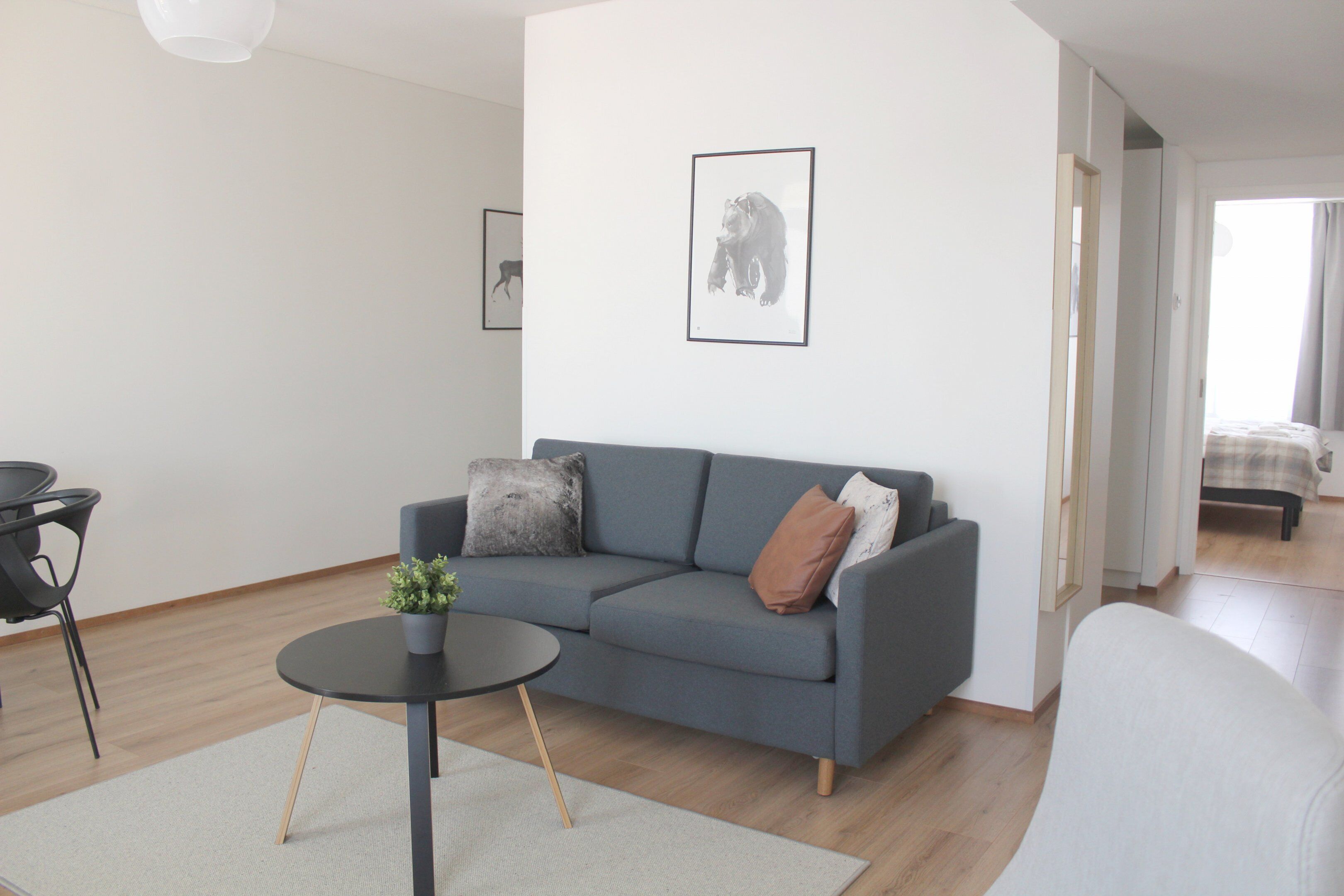 Un salon moderne avec un canapé gris, une petite table ronde ornée d’une plante et un tableau encadré accroché au mur.