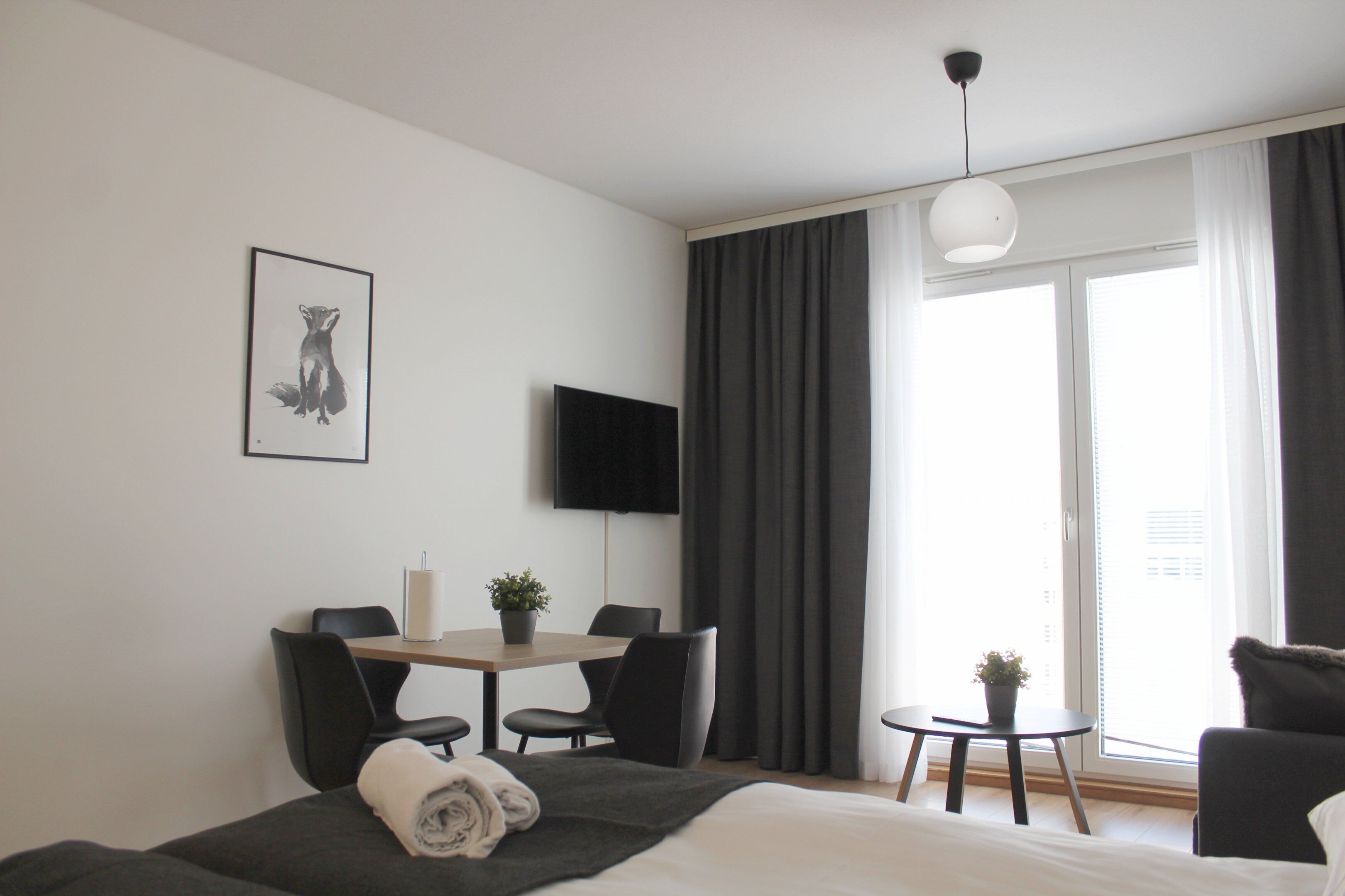 A modern hotel room with a bed, a desk with chairs, a small table with a plant, a flat-screen TV, and a large window with curtains.