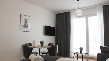 A modern hotel room with a bed, a desk with chairs, a small table with a plant, a flat-screen TV, and a large window with curtains.