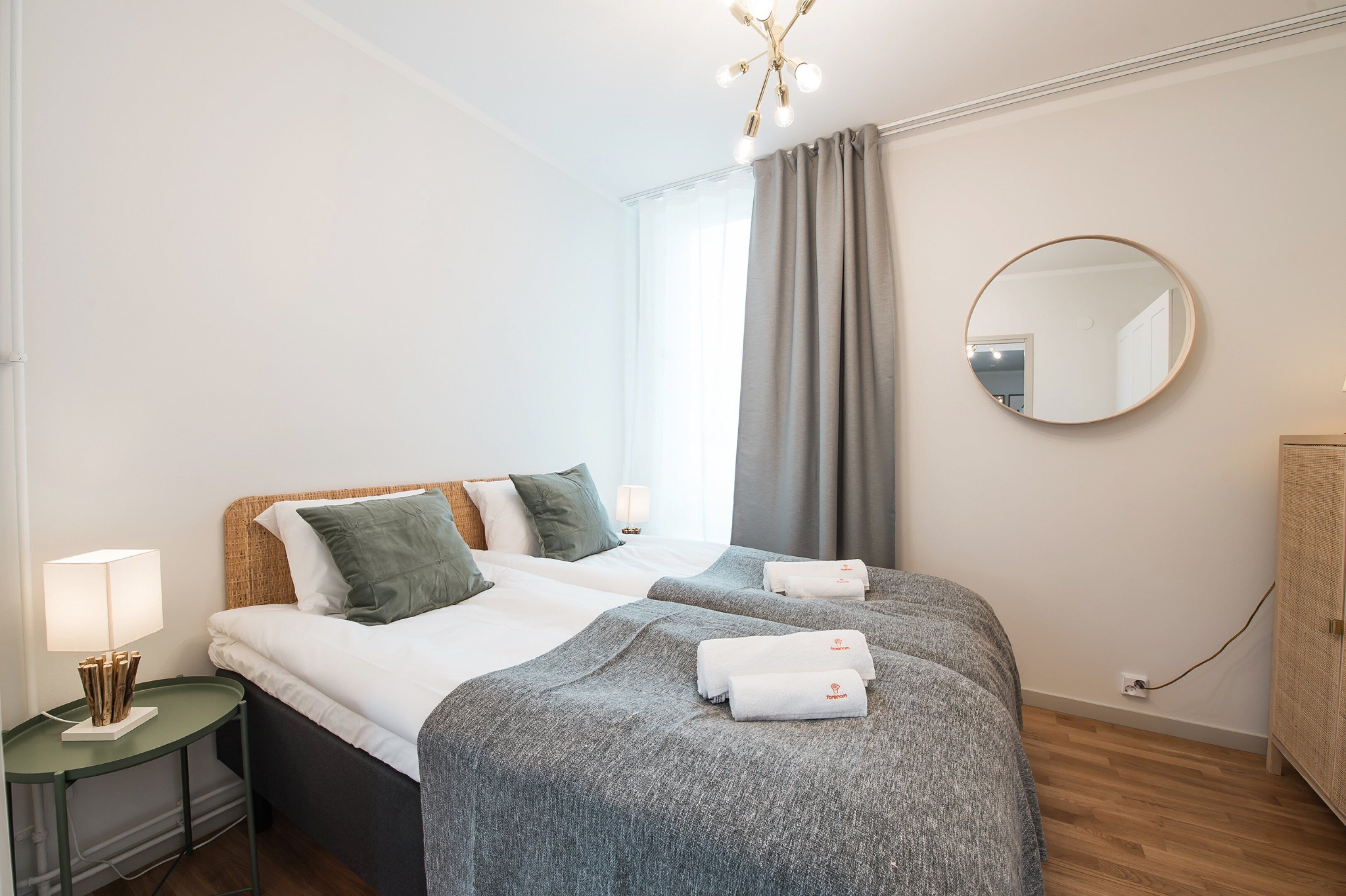 A hotel room with two beds, a round mirror, a nightstand with a lamp, and a wardrobe.