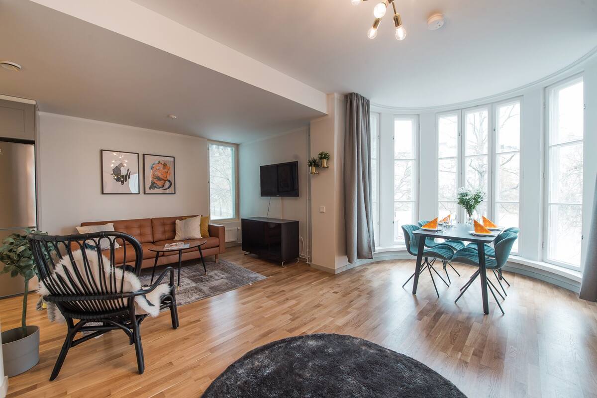 Un salon spacieux doté d’un plancher en bois, d’un fauteuil noir, d’un canapé marron, d’une table à manger avec des chaises et d’une grande fenêtre avec des rideaux.