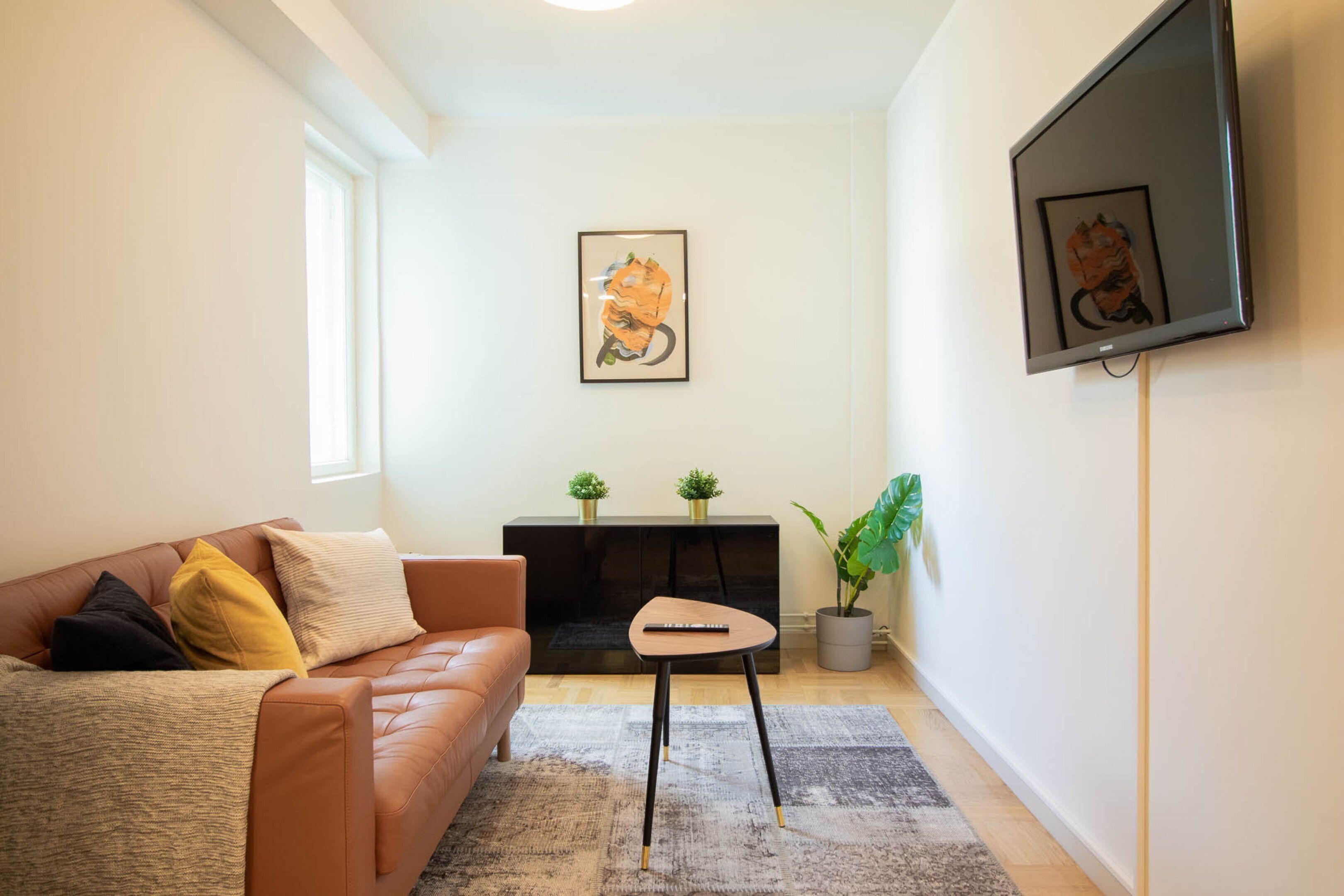 Un salon avec un canapé en cuir marron, une petite table en bois, un meuble de rangement noir et un grand téléviseur fixé au mur.