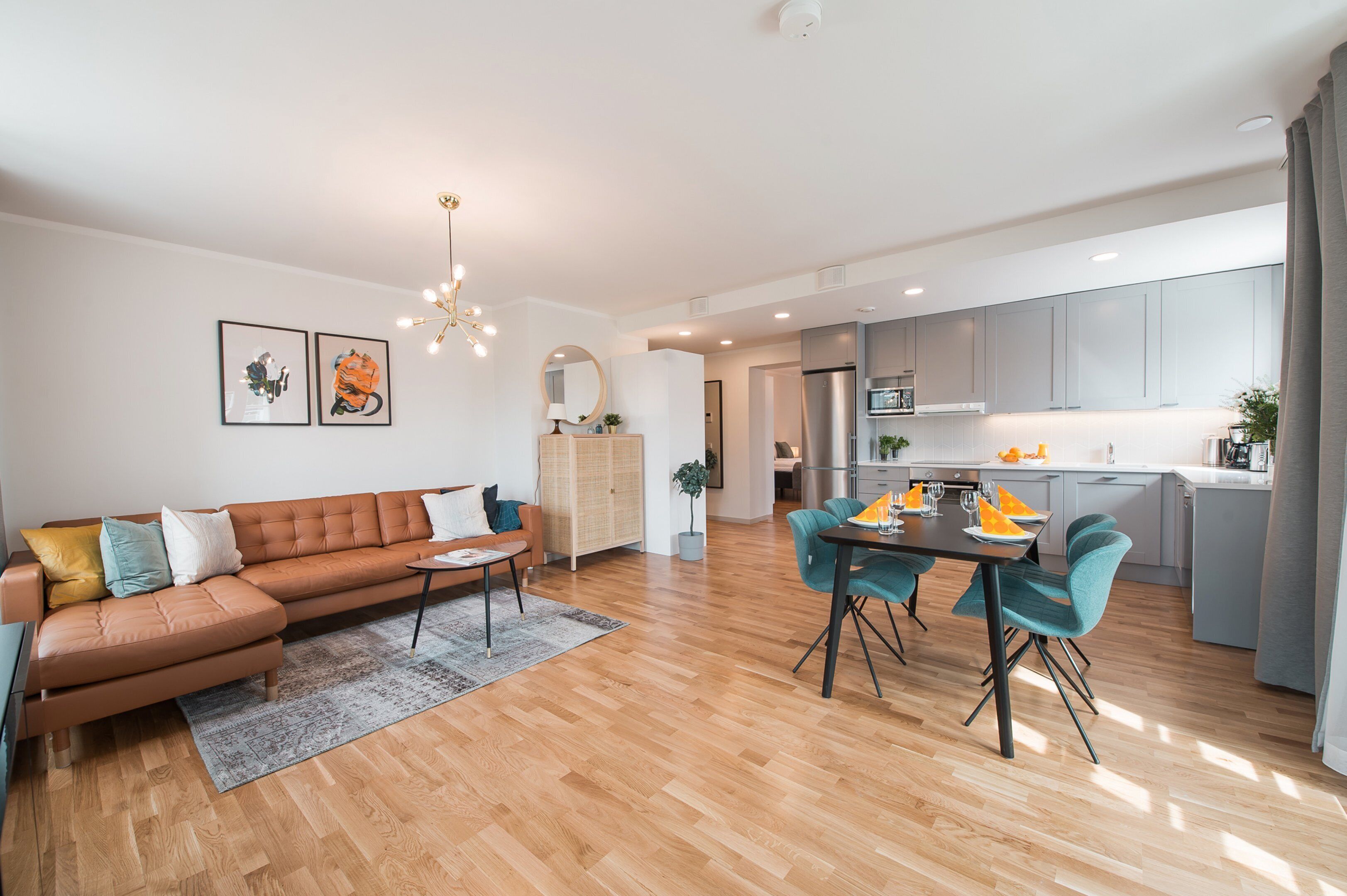 A modern living room with a sofa, dining table, and chairs, connected to a kitchen area with grey cabinets and a stainless steel refrigerator.