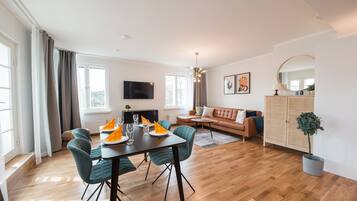 A modern living room with a dining area, wooden flooring, a sofa, a TV, and a large window.