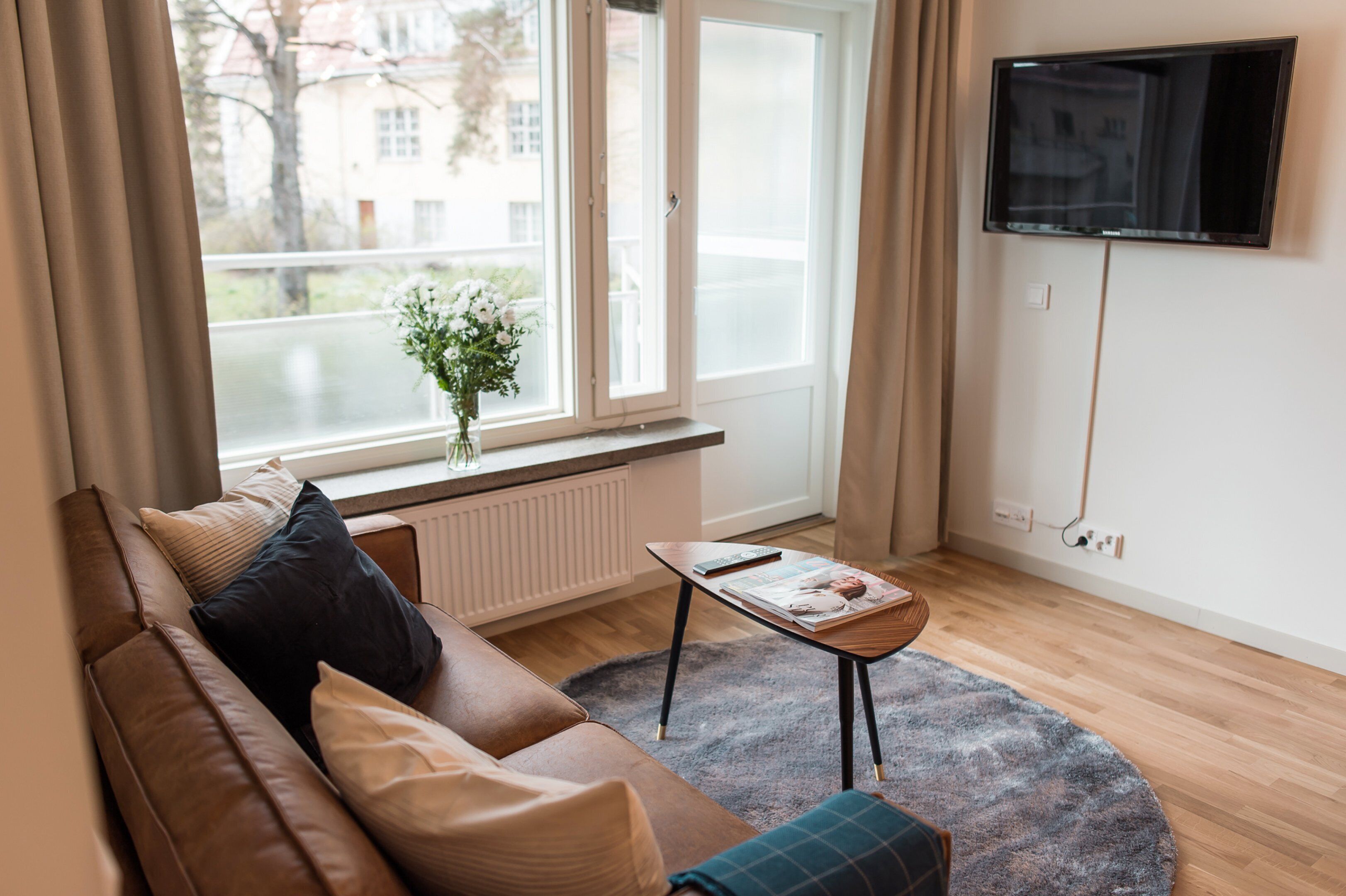 A modern living room with a brown leather sofa, a small round table with a magazine, a TV mounted on the wall, and a window with a view of buildings and trees.
