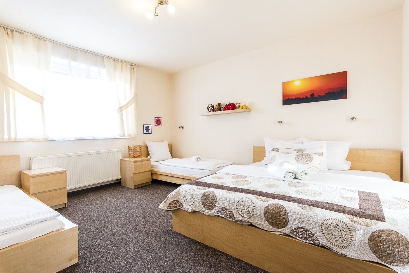 A well-lit bedroom with two beds, a radiator, a window with curtains, a shelf with decorative items, and a sunset painting on the wall.