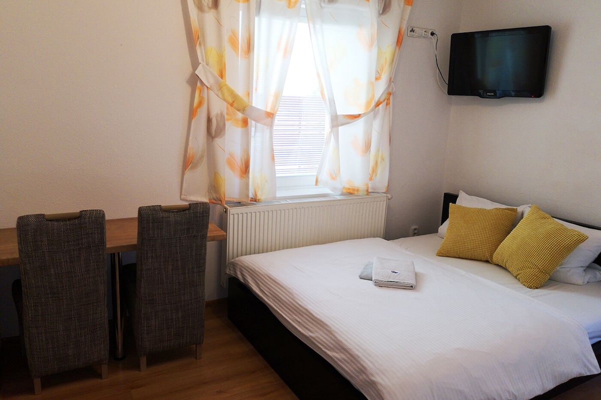 Habitación de hotel con cama, escritorio con dos sillas, televisor montado en la pared y ventana con cortinas estampadas.