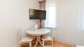 Una habitación pequeña y limpia con una mesa redonda y dos sillas, un televisor de pantalla plana montado en la pared y una ventana con cortinas traslúcidas.