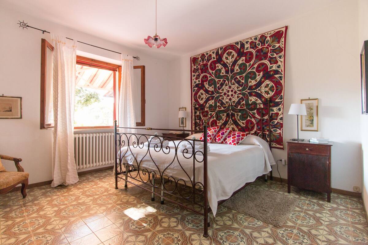 Um quarto com cama, radiador, janela com cortinas, tapeçaria estampada e uma pequena mesa de cabeceira.