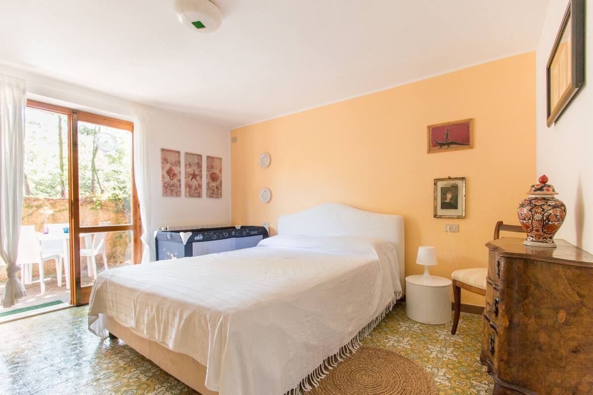 Quarto com cama, mesa pequena, cadeira, cômoda e janela com cortinas.