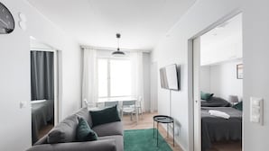 A modern, well-lit room with a grey sofa, a small round table, a bed with dark bedding, and a TV on the wall. - Forenom Serviced Apartments Neilikkatie (Vantaa)