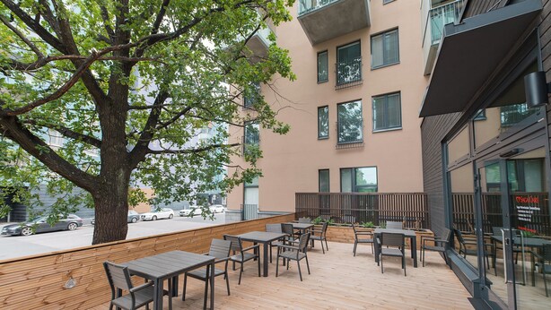 An outdoor seating area with tables and chairs, a large tree, and a building with multiple windows and balconies. - Forenom Serviced Apartments Neilikkatie (Vantaa)