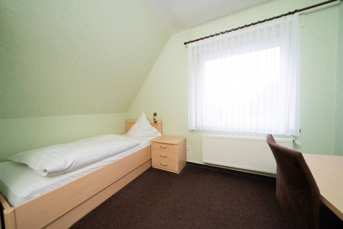 A small, single-bed room with a wooden bed, a nightstand, a chair, and a window with sheer curtains.