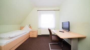 A small, neatly arranged room with a single bed, a desk with a chair, a television, and a window with curtains.