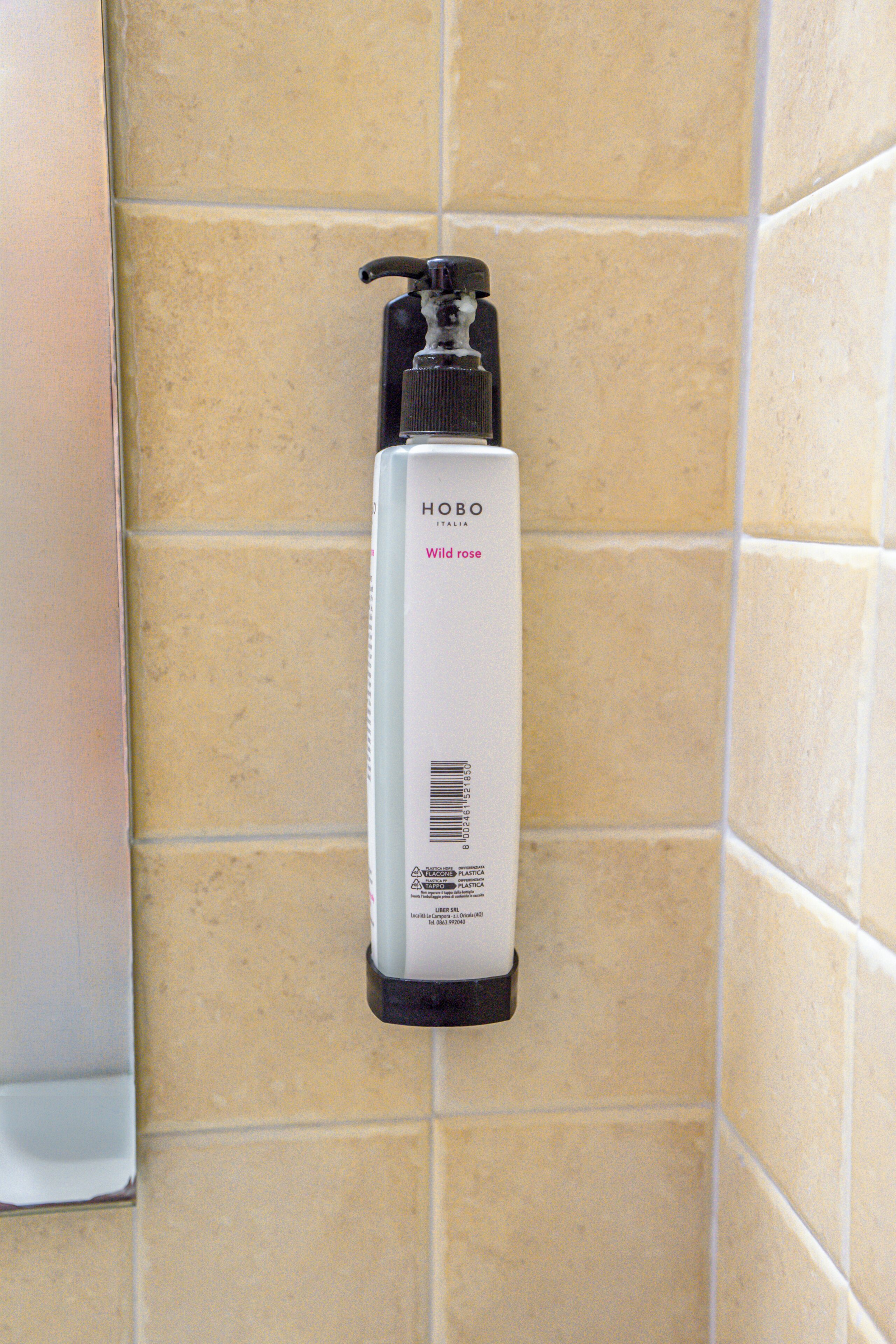 Bany amb rajoles beix i un dispensador de xampú HOBO Wild rose de color blanc fixat a la paret.