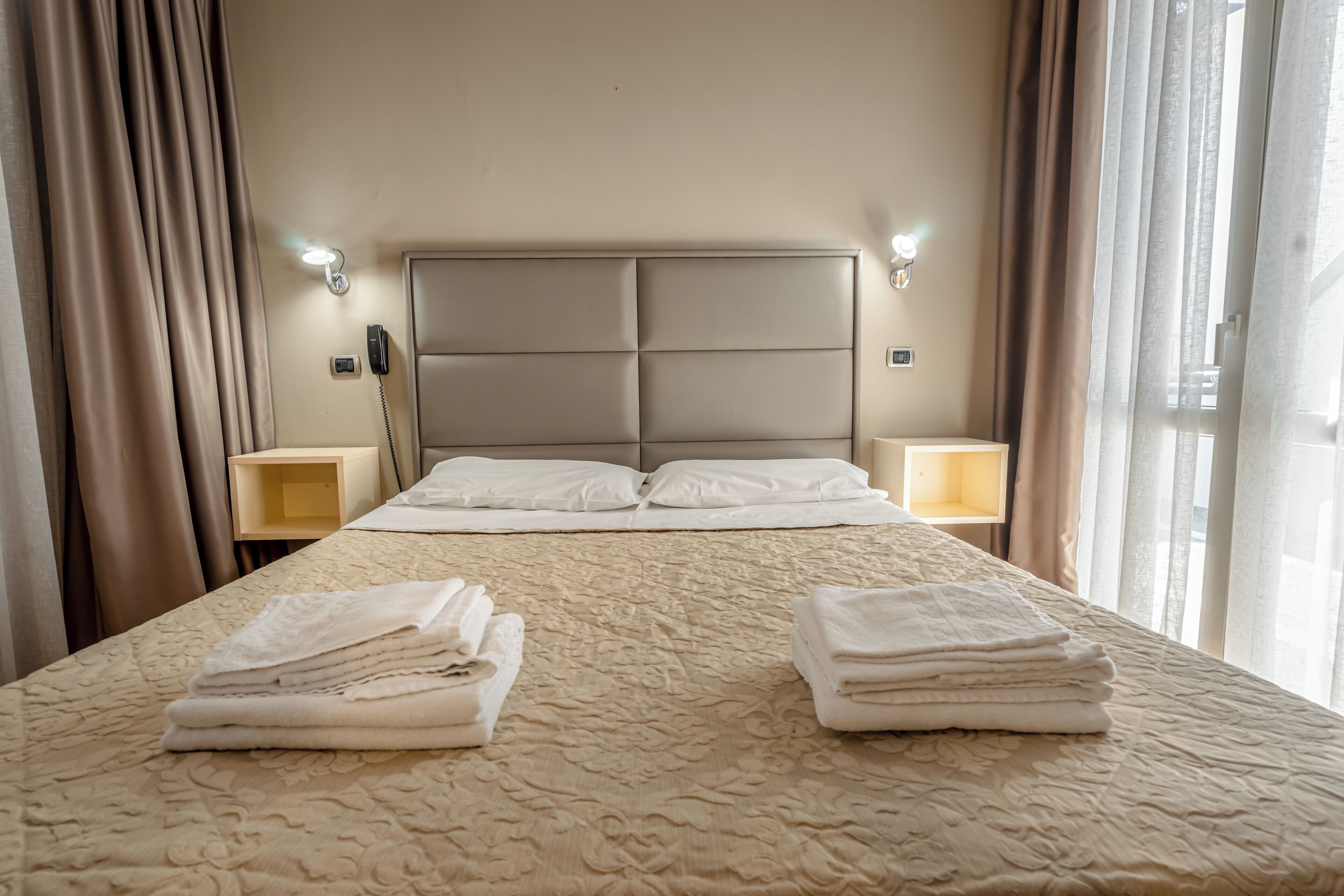 Habitación de hotel con una cama grande, dos mesitas de noche, cabecero y una ventana con cortinas.