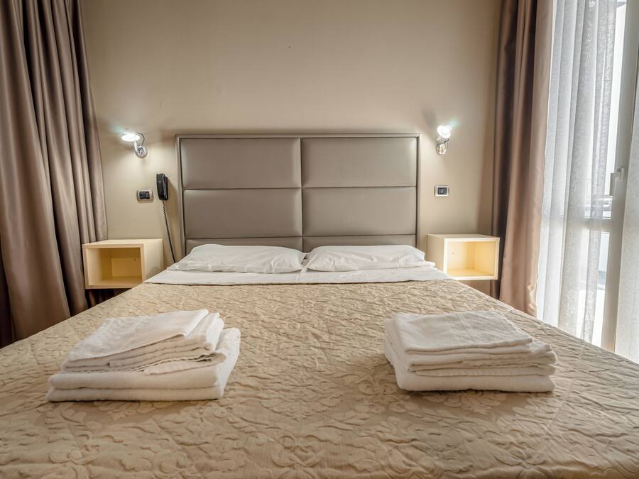 Habitación de hotel con una cama grande, dos mesitas de noche, cabecero y una ventana con cortinas.